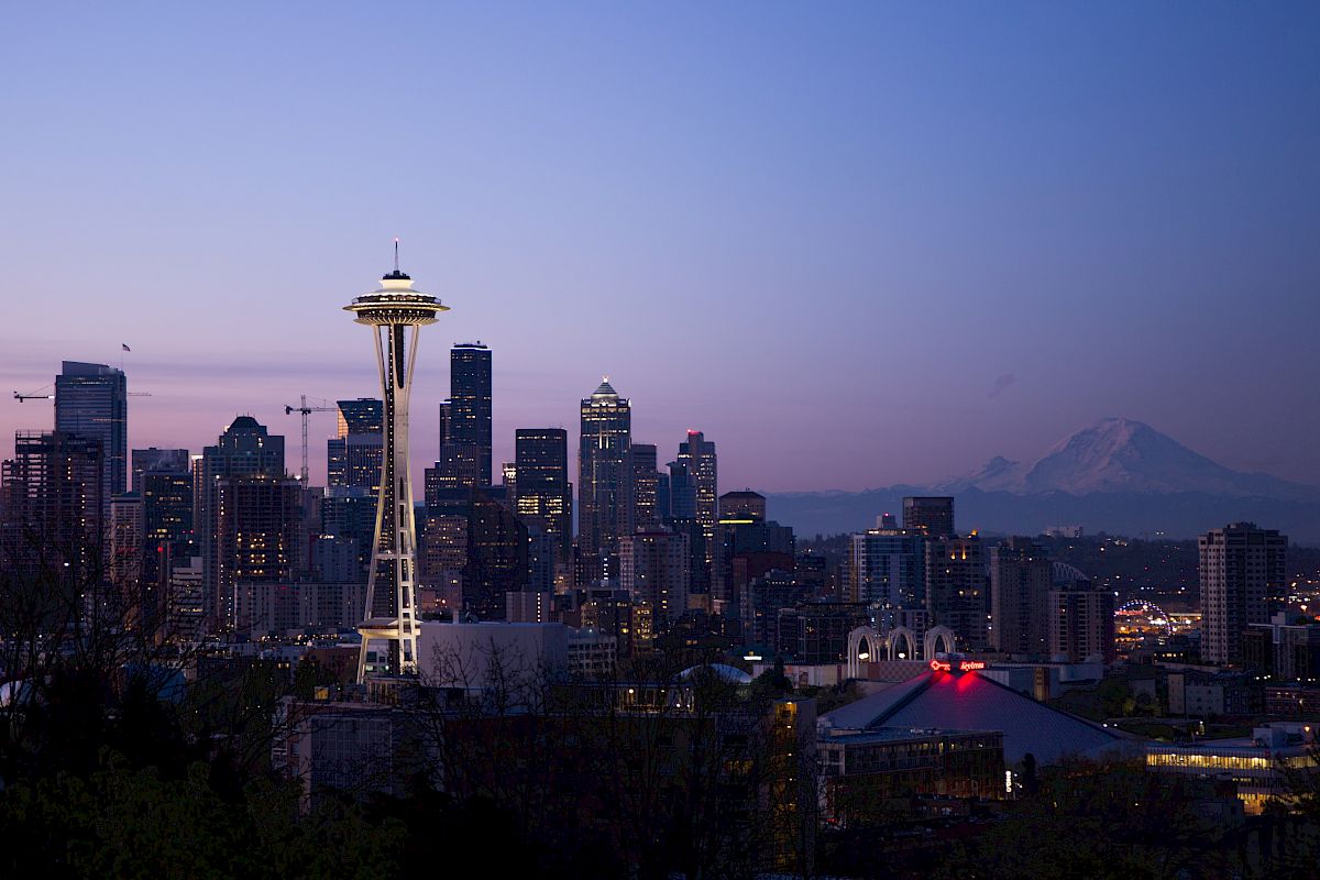 This image shows the skyline of Seattle at dusk, featuring the Space Needle prominently and Mount Rainier in the background against the fading light.