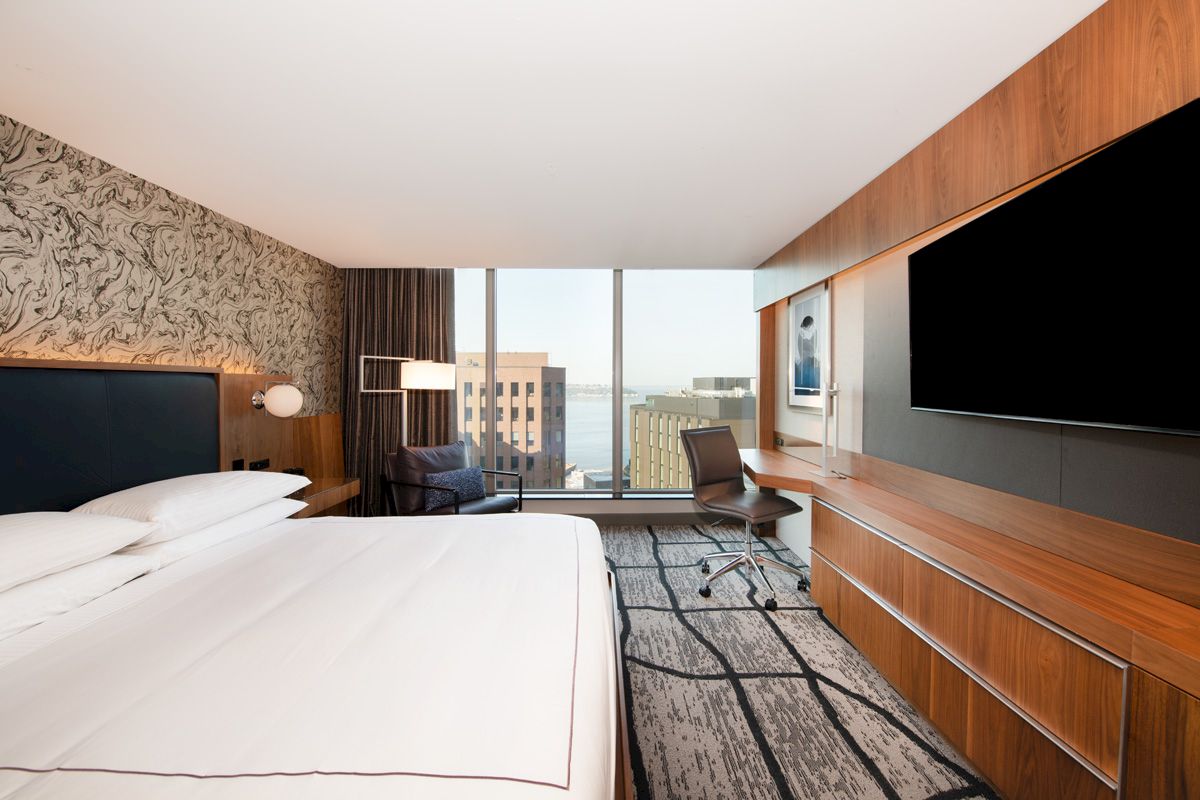 A modern hotel room with a large bed, flat-screen TV, desk, chair, and city view through large windows.