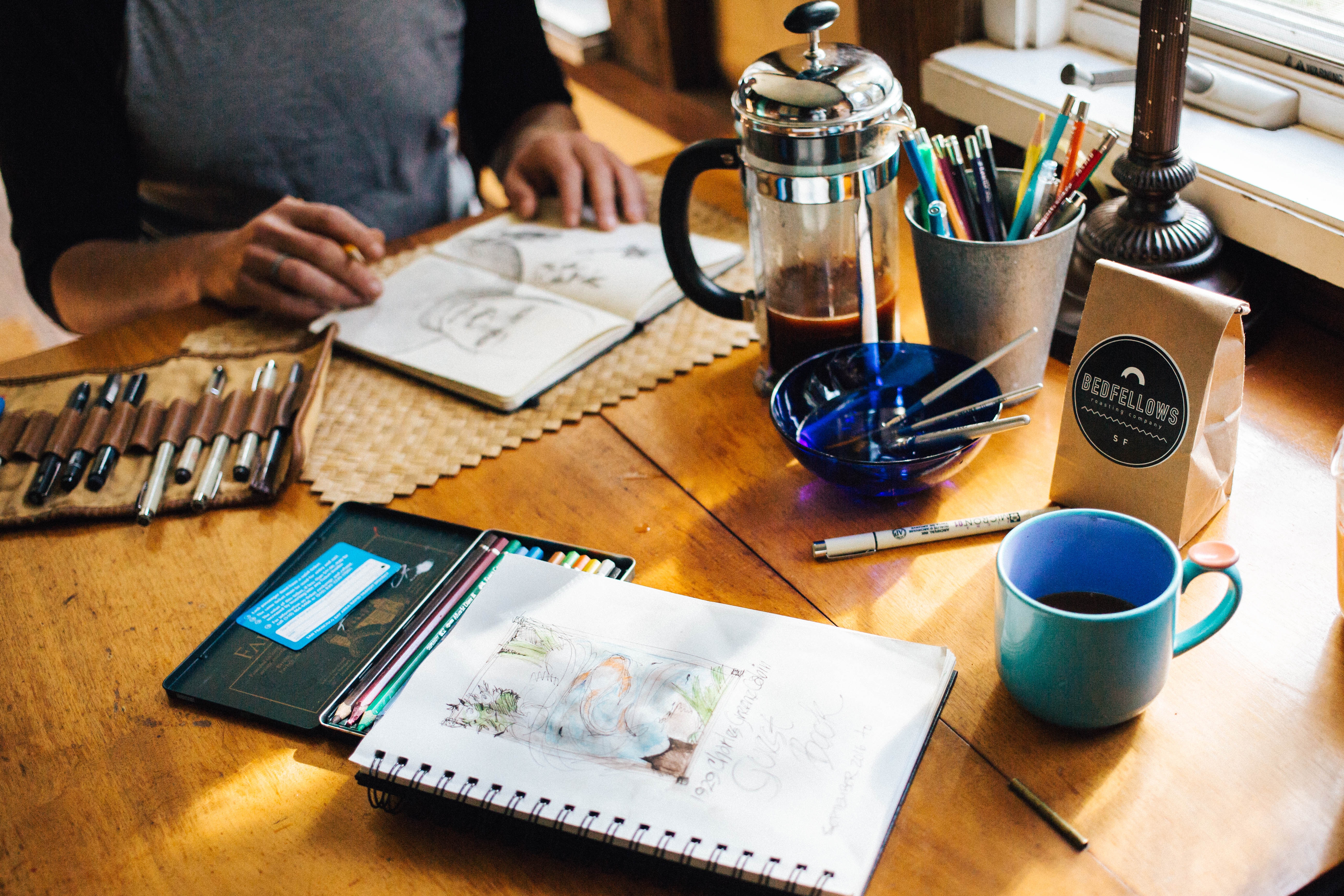 A person is sketching at a table with drawing supplies, a French press, coffee, and a bag of coffee beans. There is also a notebook on the table.