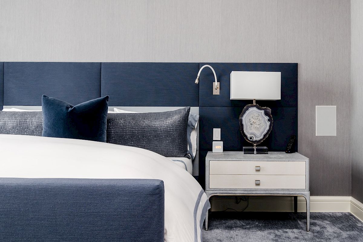 A modern bedroom features a bed with blue elements, white linens, a nightstand with a lamp, and stylish decor on a carpeted floor.